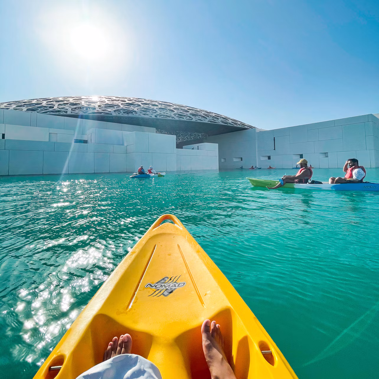 People kayaking in clear turquoise water near a modern white building with a domed roof under a bright sun.