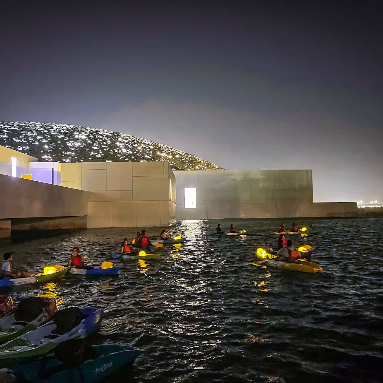People kayaking in a dark, illuminated water area near a modern building with a latticed, dome-shaped roof in the background.