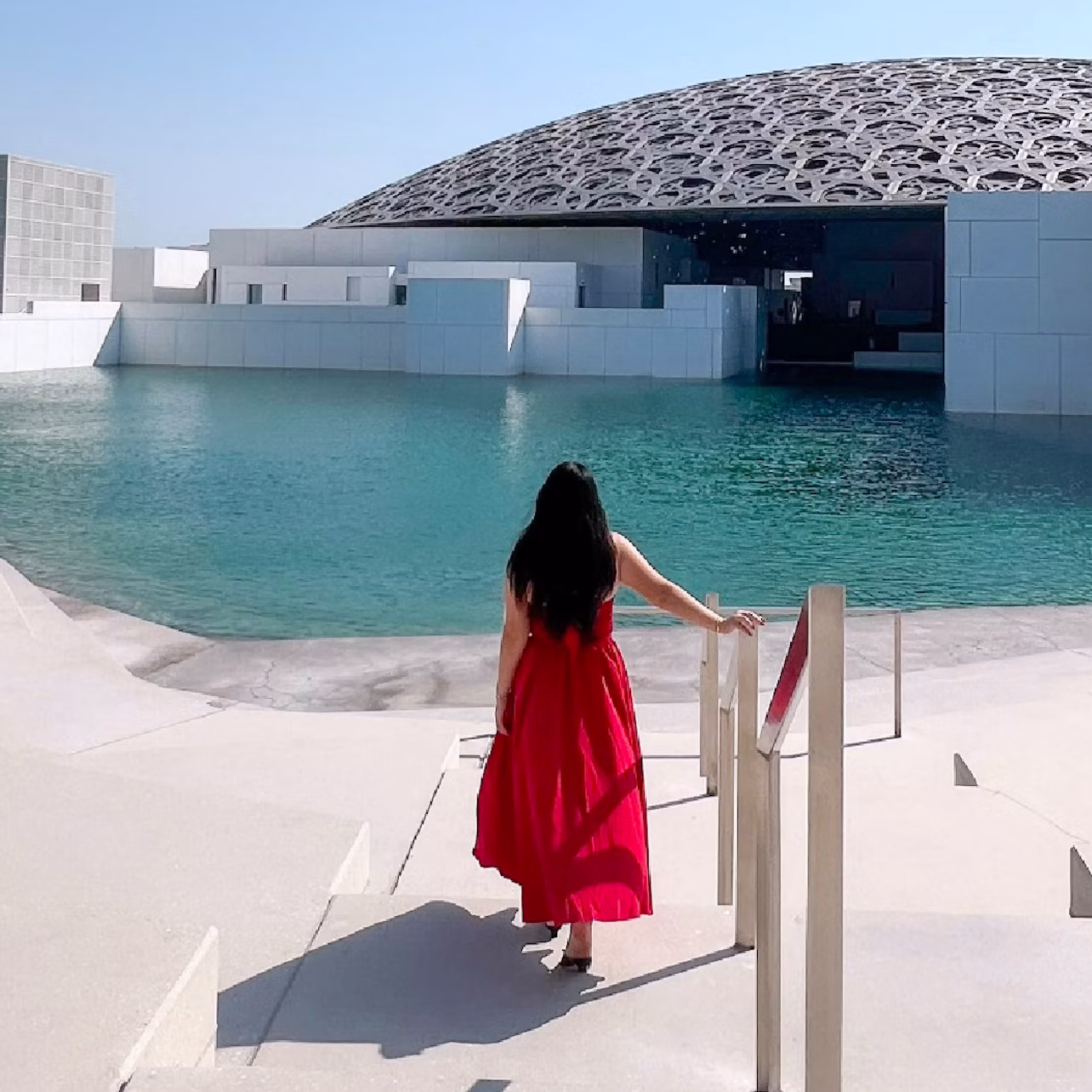 A woman in a red dress stands near a railing, overlooking a water feature and a domed building under a clear blue sky.