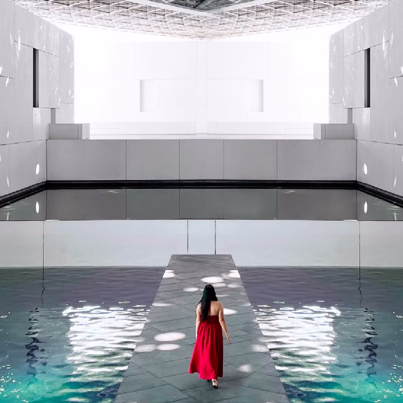 A woman in a red dress walks on a narrow path surrounded by water, inside a modern, white architectural space.