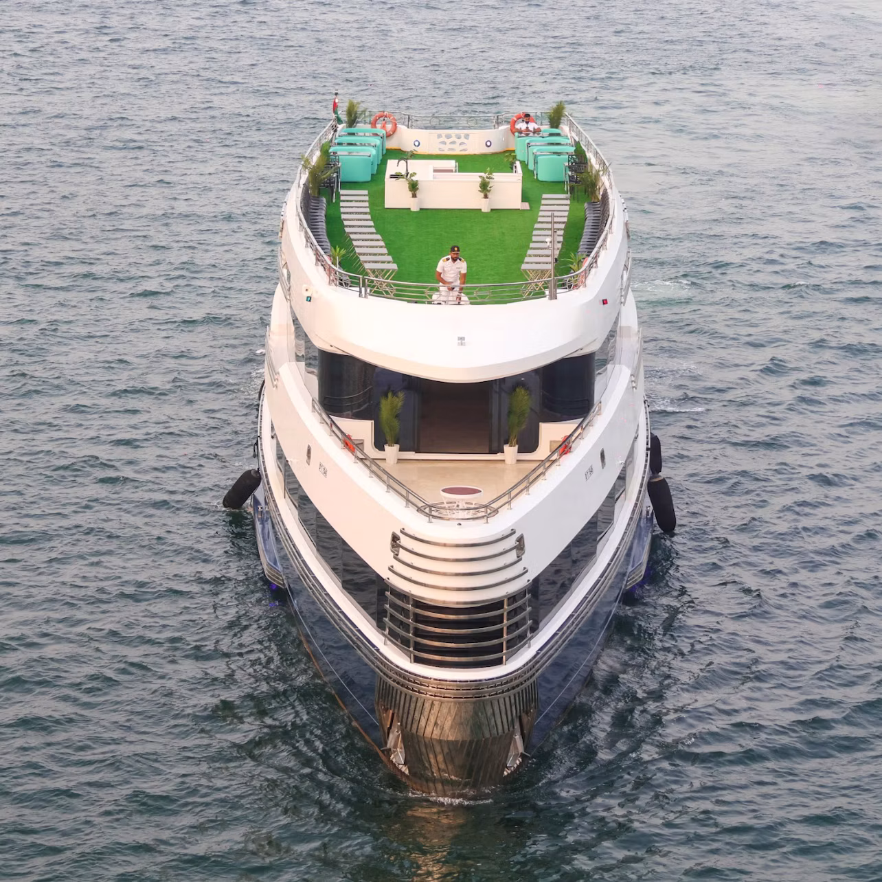 Front view of a luxury yacht with multiple decks, green turf, and a couple standing on the upper deck, surrounded by water.