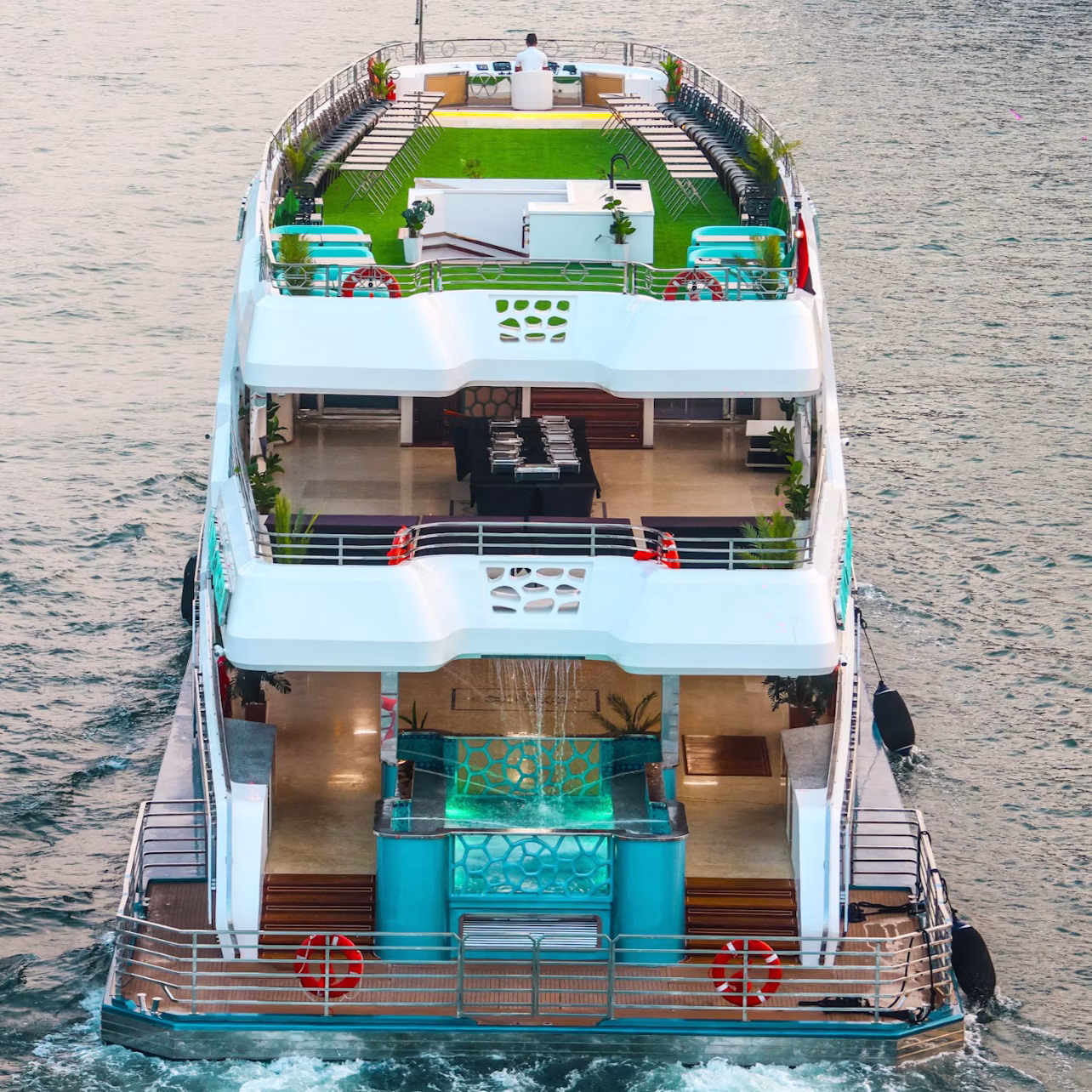 A multi-level yacht with a green rooftop deck, outdoor seating, and a modern design cruising on the water.