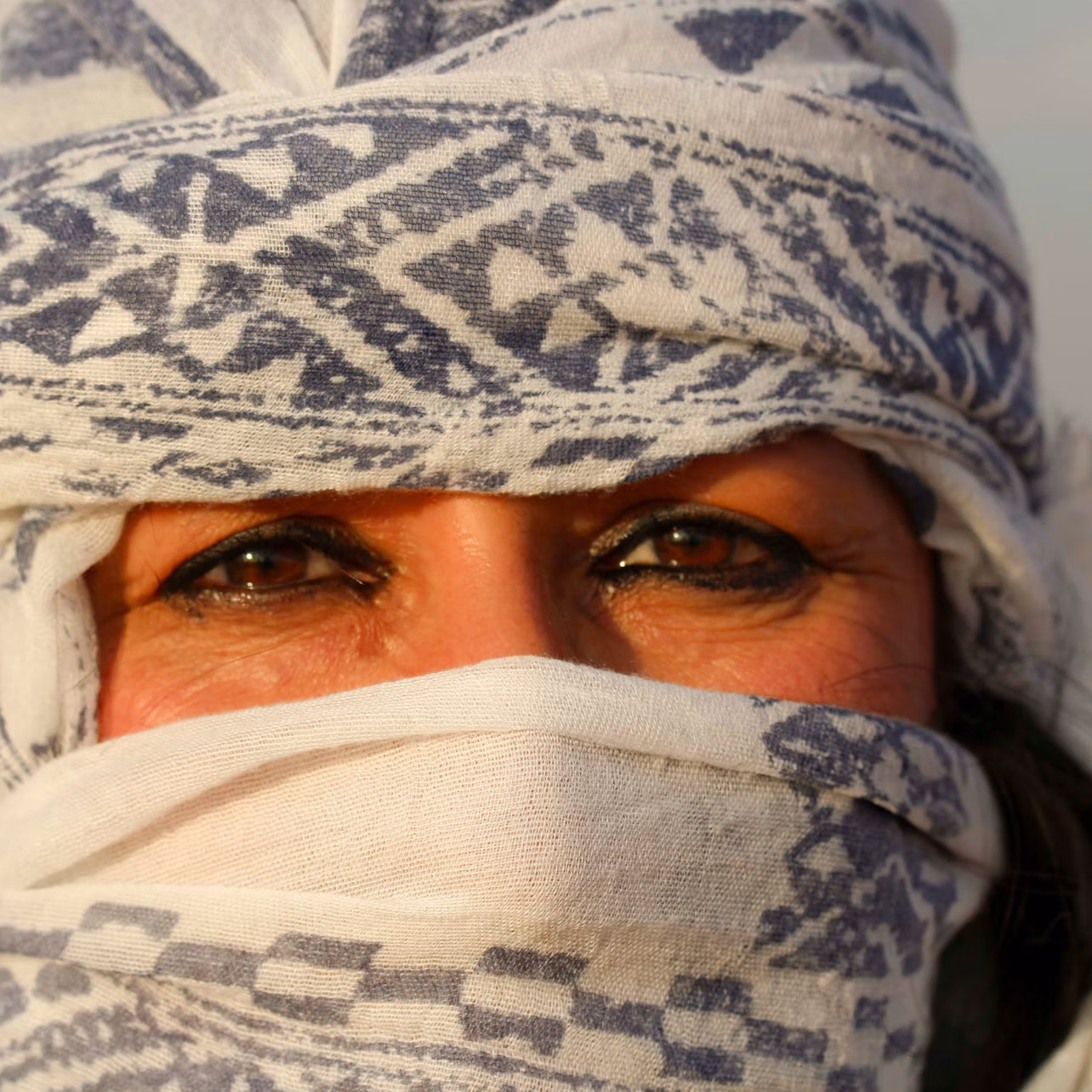 Close-up of a person's face with deep brown eyes, partially covered by a patterned white and blue cloth.