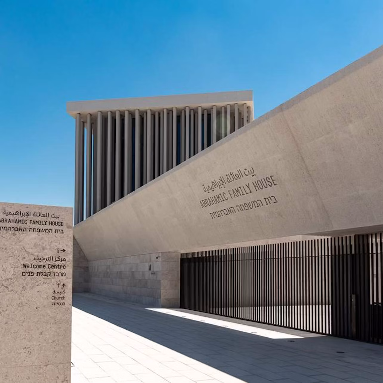 Modern building with the text "Abrahamic Family House" inscribed on an angled wall. Vertical columns and a blue sky are visible.