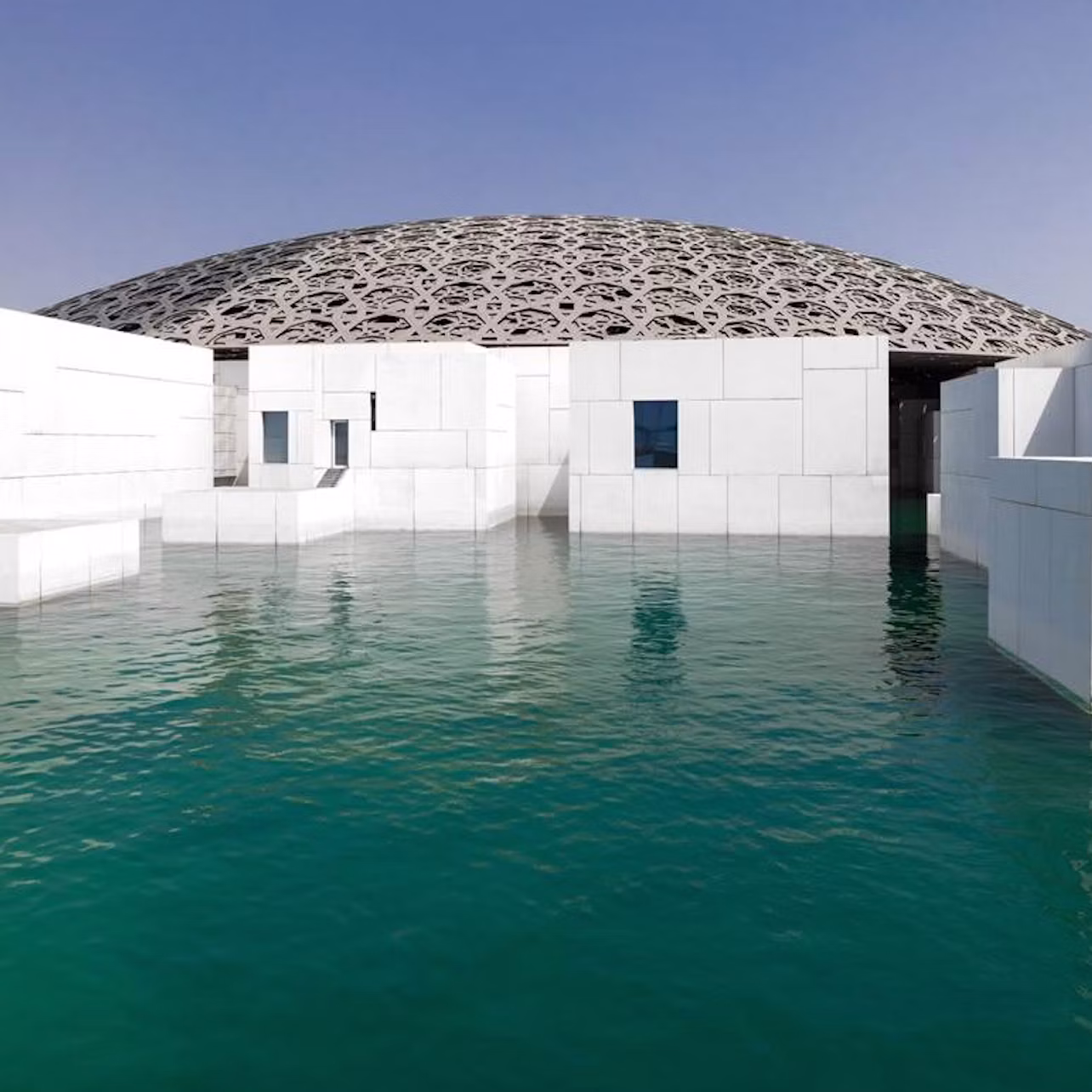 Modern white building with large, intricate dome roof, surrounded by turquoise water under a clear blue sky.