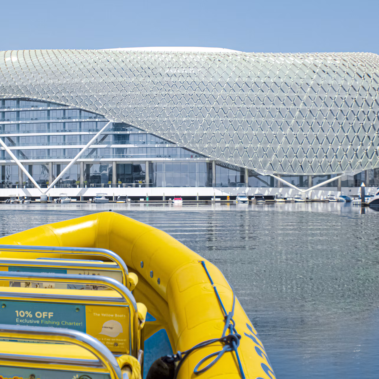 Yellow boat on water faces a modern building with a lattice structure. Floating docks with boats are visible in the background.