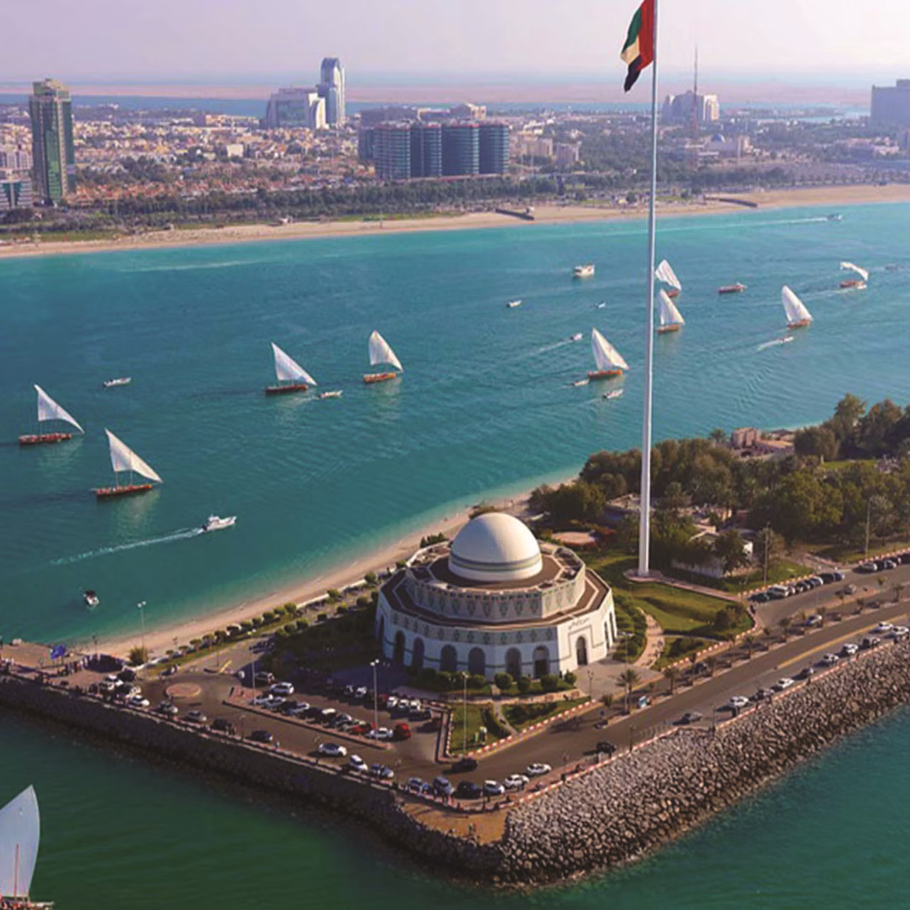 Aerial view of a waterfront city with a domed building, large flagpole, and numerous sailboats on the water.