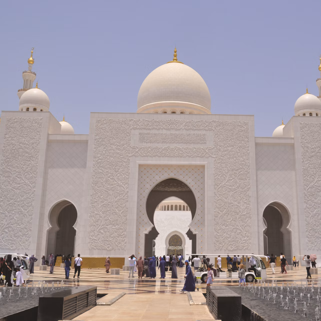 Large white mosque with intricate designs, gold accents, and multiple domes. People walk around the courtyard under a clear sky.