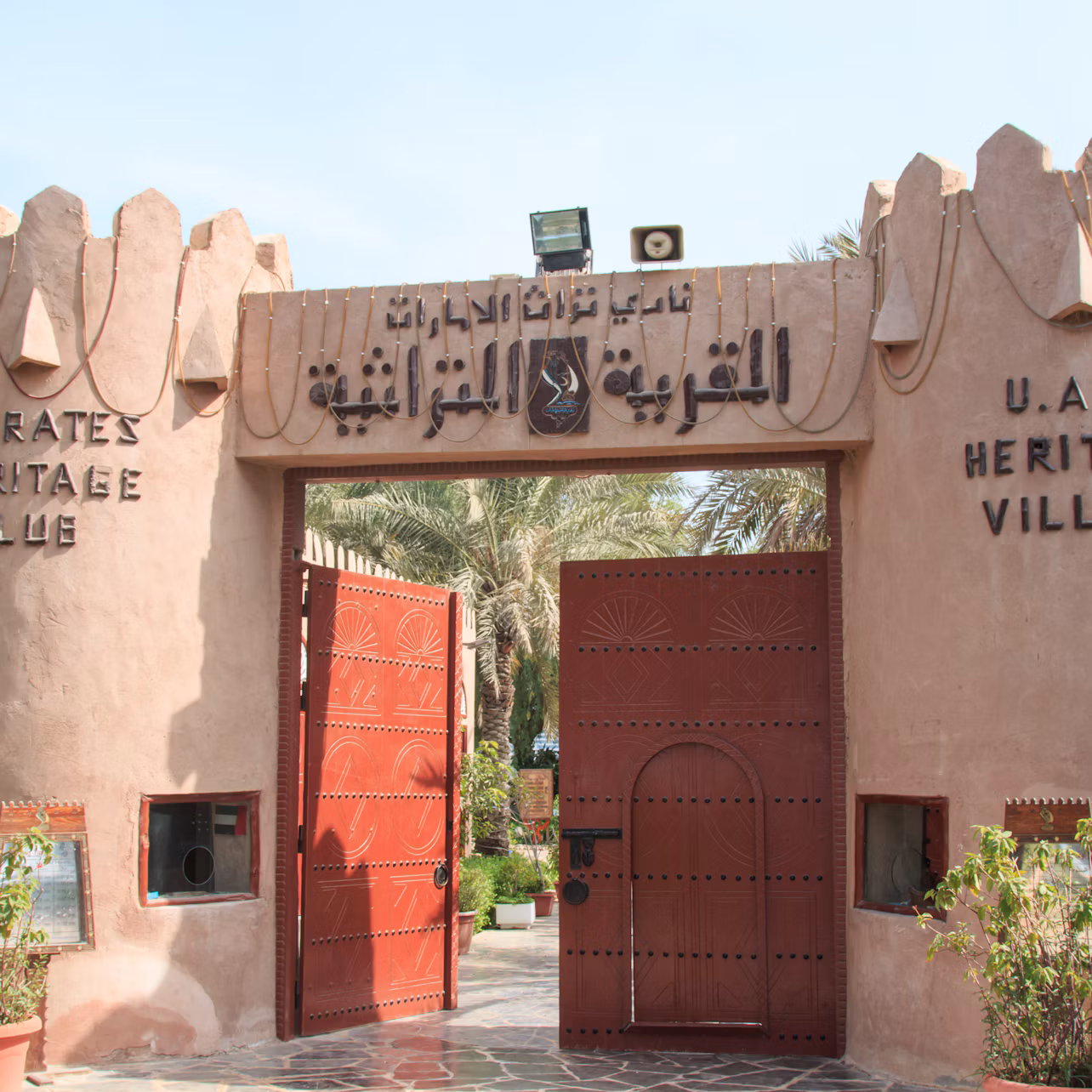 Entrance to the UAE Heritage Village with traditional-style gates and Arabic and English text above. Trees and plants are visible inside.